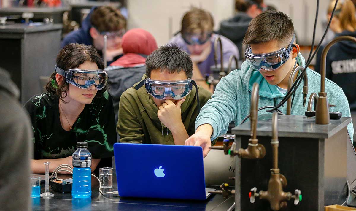 A group of students in a lab, wearing goggles and looking at a computer.