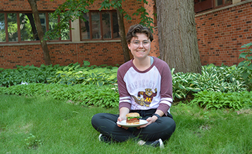 Maddi Johnson has short brown hair and fair skin, she sits cross legged in green grass holding a plate with a plant-based burger on it. 
