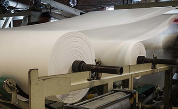 Large sheets of paper spanning an expansive factury floor spin onto a gian roll in a paper manufacturing company. 