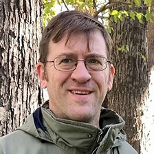 Headshot of Chris Lenhart wearing glasses and a dark green jacket posed in-front of a tree.