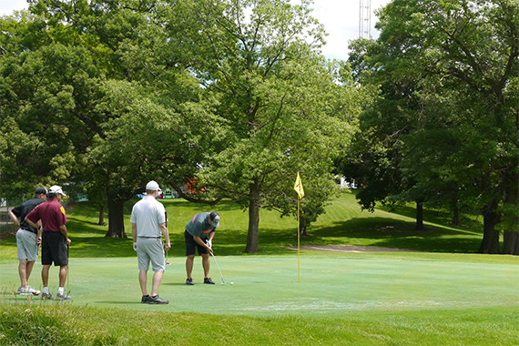 Three golfers watch as a fourth puts his golf ball into the course hole. 