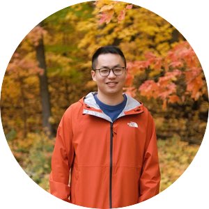Jie Yang smiling and wearing a windbreaker. Posed outdoors in front of bright fall leaves