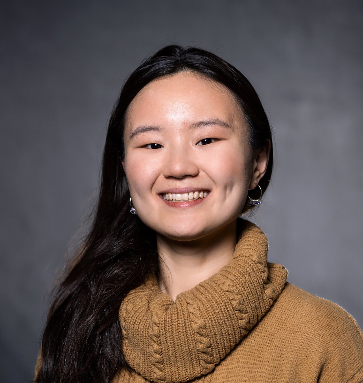 Rui Cheng in a professional headshot smiles with her long black hair pulled in front of her right shoulder, wearing a gold turtleneck knitted sweater