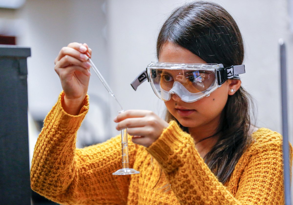 A student wearing protective goggles concentrates on using an eyedropper to pour liquid into a test tube.