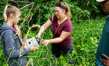 Undergraduate students stand in a field of tall grass not far from lake shore while they work together to get a water collecting device turned on to take well water samples.