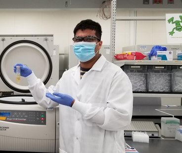 Raghav works in a lab wearing a white coat, safety goggles, and gloves while holding up a vile with yellow fluid in it. 