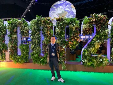 Dr. Roger Raun standing in-front of a colorful sign covered in plants that reads "COP26". Further behind him is a large projected image of earth.