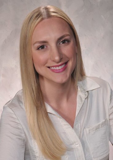 Professional headshot of Megan Acker who has long blonde hair and is posed in front of a grey background.