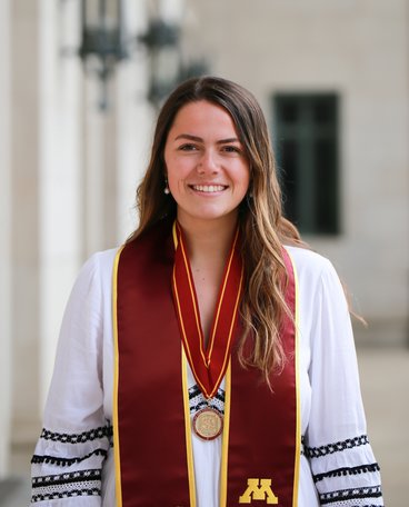 Sarah Whiteside has long brown hair and is wearing a white top with black embroidery with a maroon U of M honors stole for graduation.
