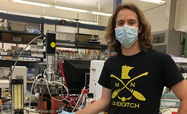 Lief van Lierop stands in front of a lab setup wearing a blue surgical mask and a black tee shirt.
