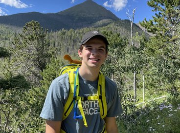 Kyle has short brown hair and wear a baseball cap as he hikes along a trail posing in front of a mountain.