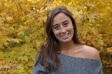 Katie has long brown hair, freckles, and blue eyes and is posed in front of yellow fall leaves.