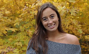 Katie has long brown hair, freckles, and blue eyes and is posed in front of yellow fall leaves.
