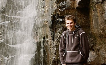 Alex Hilde has gelled brown hair and smiles as he stands in front of a waterfall wearing a gray hoodie. 