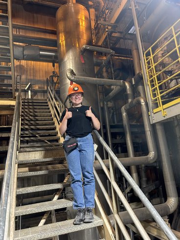 Eilee Keske poses on a stairwell in the Sappi paper mill wearing an orange hard hat with a black t-shirt and jeans.