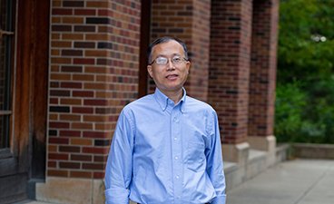 Hua Zhao poses for a head shot in front of a brick building on the St. Paul campus, wearing a blue button up shirt. 