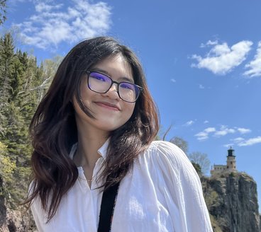 Julie wears black rim glasses and has long dark brown hair, wearing a white shirt as she poses in front of Split Rock Lighthouse on the north shore of Lake Superior.