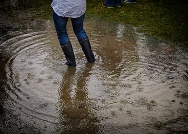A person stands in ankle deep water wearing rain boots. 