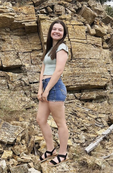 Brooke poses in front of a natural stone cliff wearing a cropped tee and jean shorts.