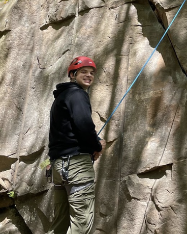 Kyle stands in a climbing harness and helmet holding a climbing rope along a cliff outside.