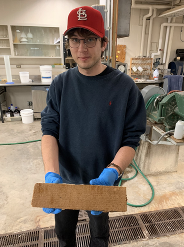 Max wears a red St Louis baseball cap and holds a piece of composite wood in Kaufert lab.