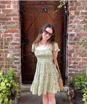 Megan stands in front of a wood door and brick wall wearing a light green dress with white flowers.
